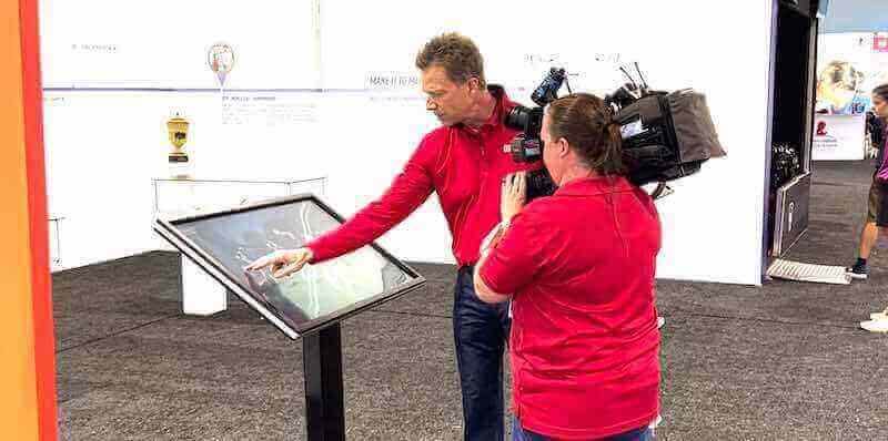 Interactive touchscreen kiosk being demonstrated — commercial-grade hardware and purpose-built software are both required for installations that will run without issues for years Camera operator filming a man demonstrating an interactive touchscreen kiosk at a public exhibit