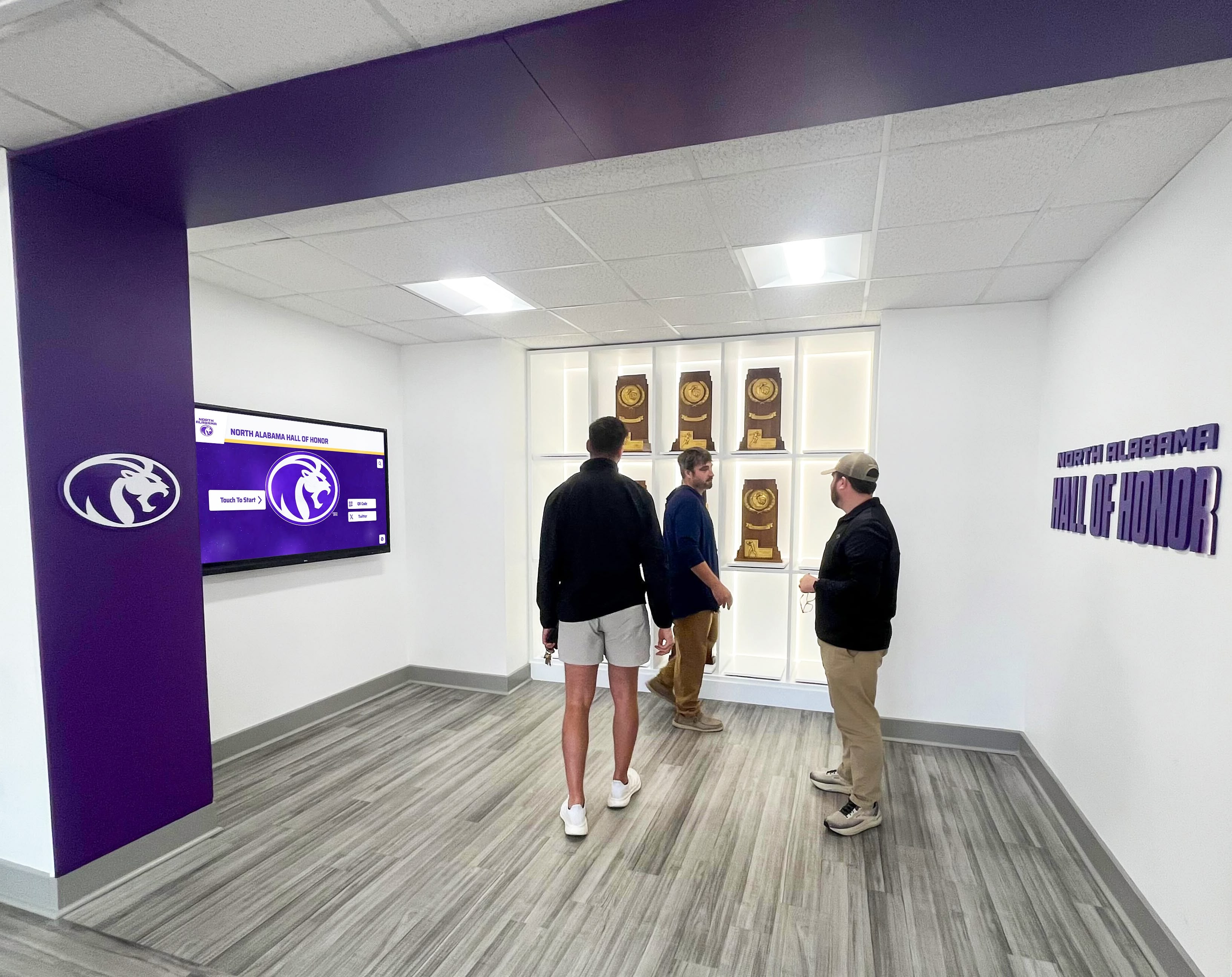 Three men inside North Alabama Hall of Honor exploring trophy display and athletic recognition wall